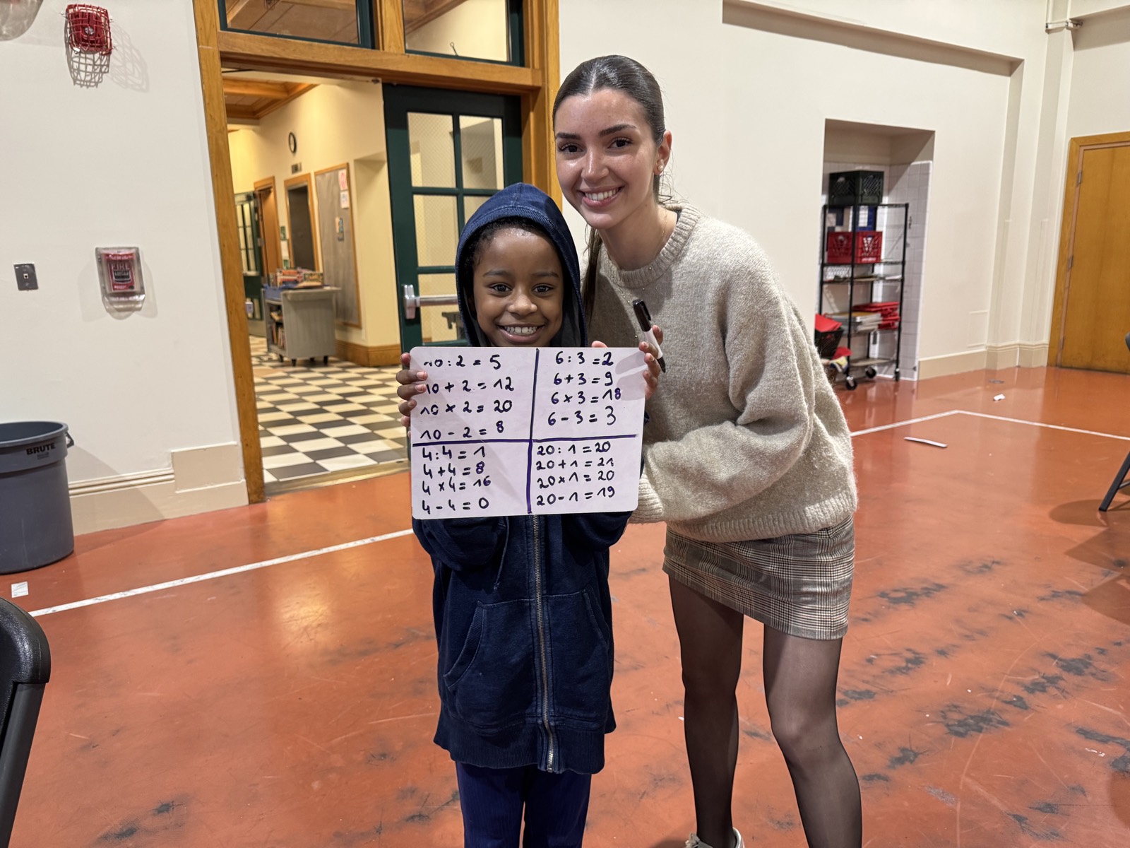 A child proudly holding up a whiteboard of finished math problems beside a volunteer